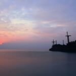 silhouette of person standing on rock formation in the middle of sea during sunset